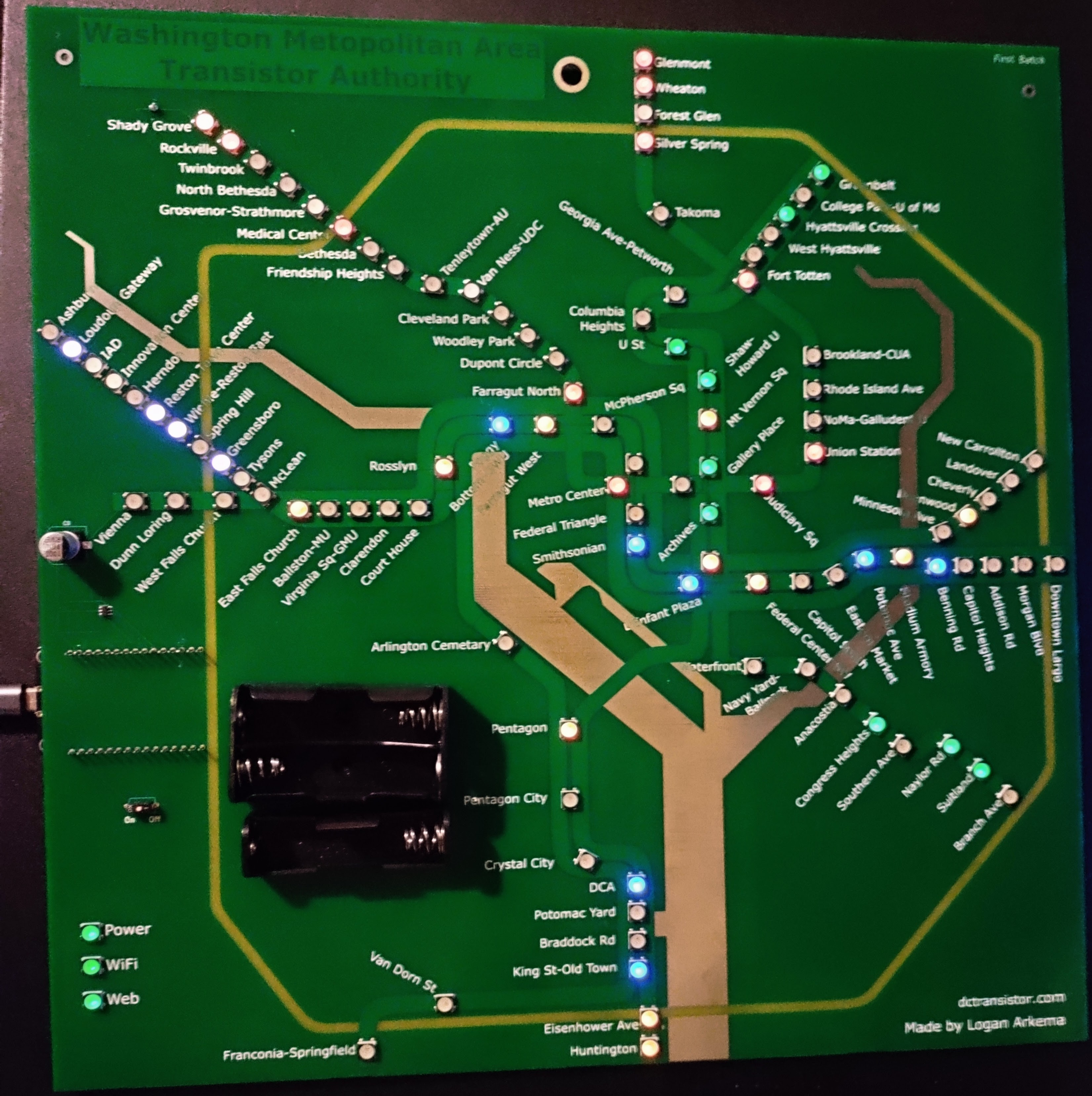 Large picture of a Development DCTransistor board, powered on to show live train positions. The board is programmable circuit board, mostly light green except for a dark green title and dark green rectangles representing the DC metro train lines, a beige boundary representing the beltway, and silver representing the Potomac and Anacostia rivers. It is designed to represent the DC Metro System map. As an older board, it has far less refined silk screen aesthetics and looks much more shoddy compared to the Bidirectional and Standard boards.