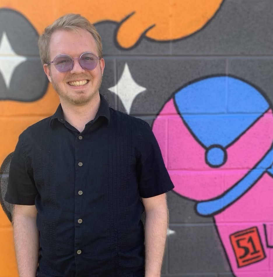 Picture of me, Logan, a young, white male with a short beard wearing purple sunglasses and black guayabera. I'm standing in front of a wall at Metrobar with art on it, to the right is a pink astronaut with a patch that says '51'